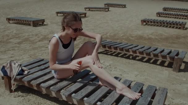 Женщина с солнцезащитным кремом наносит крем для загара на тело во время летних путешествий Женщина в отпуске с солнцезащитным кремом в пластиковой бутылке спрей, лежащей на пляже — стоковое видео