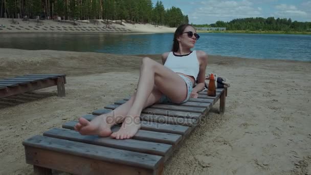 Παραλία σκηνή - κομψή γυναίκα που ακουμπά στην παραλία σε μια καλοκαιρινή μέρα στο φόντο της λίμνης στο δάσος, απολαμβάνοντας και χαμογελαστός — Αρχείο Βίντεο