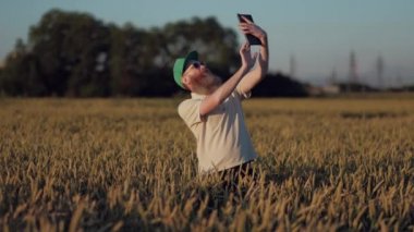 Şık adam çiftçi ile gözlük yeşil kap ve krem T-Shirt buğday dolu bir alanda bir Selfie yapıyor