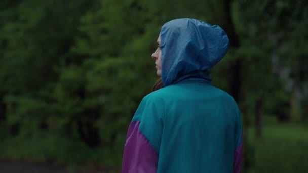 Widok - z tyłu nastolatek spacery w deszczu, w jasny kolorowy płaszcz i gumowe buty Blue usuwa kaptur z głowy — Wideo stockowe
