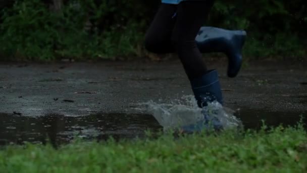 Primer plano: chica con botas de goma azules corriendo a través de los charcos, salpicando eficazmente gotas de agua, en cámara lenta — Vídeos de Stock