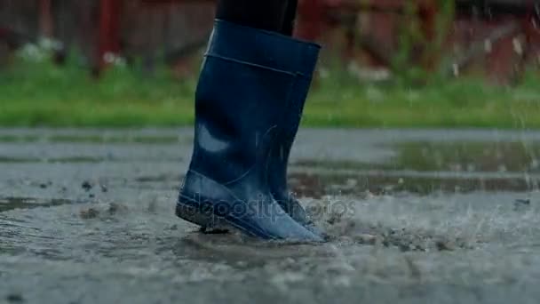 Primer plano: chica con botas de goma azules corriendo a través de los charcos, salpicando eficazmente gotas de agua, en cámara lenta — Vídeos de Stock