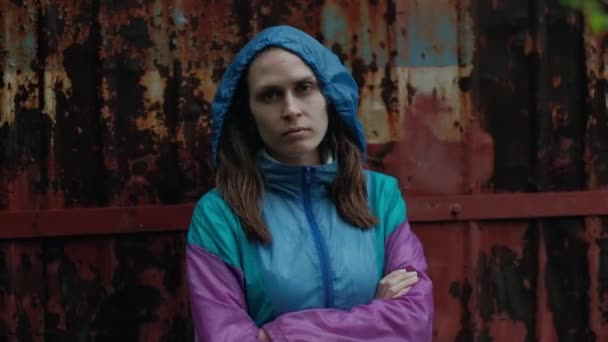 Retrato de uma mulher séria, ameaçadoramente olhando para a câmera em meio à parede enferrujada em jaqueta colorida — Vídeo de Stock
