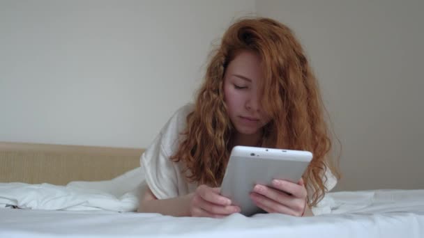 Симпатичная рыжеволосая девушка с цифровым планшетом в спальне — стоковое видео