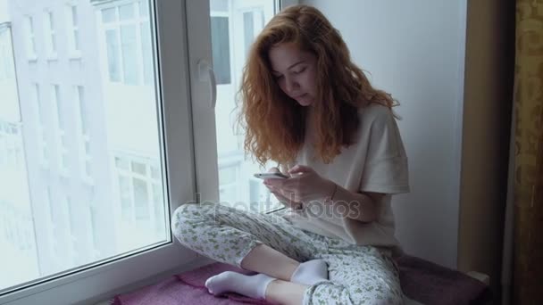 Jonge roodharige vrouw zittend op de vensterbank met een mobiele telefoon. — Stockvideo