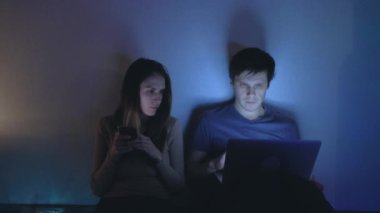 Genç çift geceleri oturup elektronik cihazlarını kullanarak iletişim kuruyor..