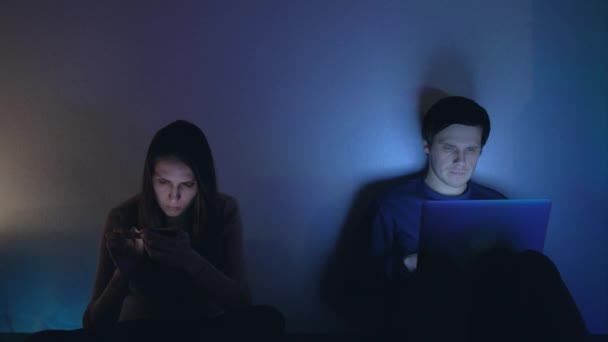 Скучная молодая пара, использующая смартфон и ноутбук ночью — стоковое видео