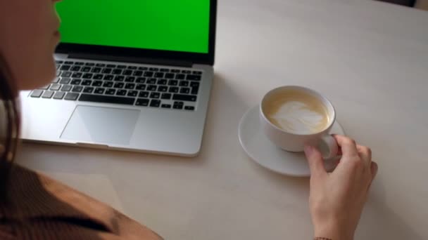 Widok z tyłu - kobieta siedząca przy stole pijąca kawę i patrząca na laptopa. — Wideo stockowe