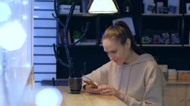 Una mujer joven usando un teléfono inteligente en un café nocturno, beber café . — Vídeos de Stock