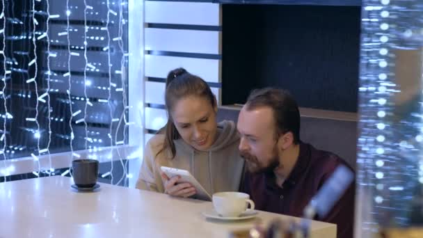 Een stel zit in een café, praat, drinkt koffie en kijkt met de smartphone. — Stockvideo