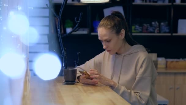 Una mujer joven usando un teléfono inteligente en un café nocturno, beber café . — Vídeo de stock