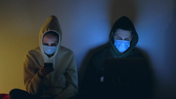 Jovem casal em máscaras médicas usando smartphone e laptop à noite . — Vídeo de Stock