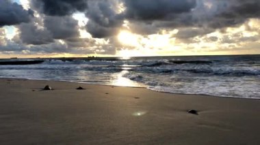 Güneş ışınları bulutları delip geçer ve denizin dalgalarını ve kumsalın ıslak kumunu aydınlatır.