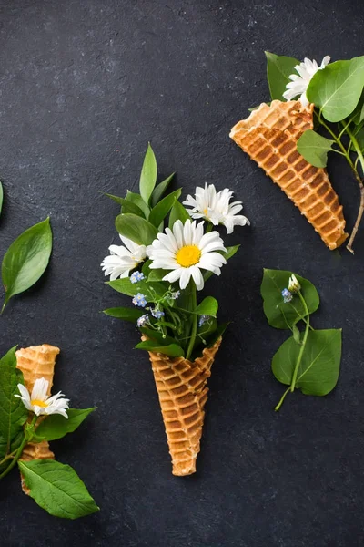 Ice cream cone with bouquet of chamomiles on a black concrete background. Fresh flowers. beautiful bouquet of wildflowers. Copy Space. Place for text. Food photo with hands. Flat lay, top view