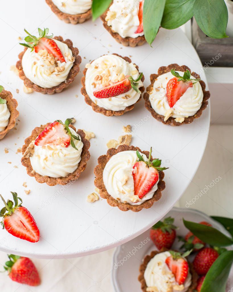 Mini tartlets with cheese cream and fresh strawberries, sprinkled with nuts on a white wooden stand. Food photo