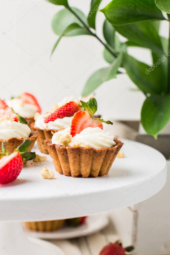 Mini tartlets with cheese cream and fresh strawberries, sprinkled with nuts on a white wooden stand. Food photo