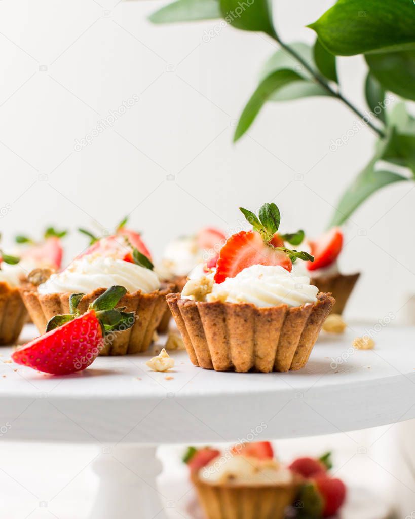Mini tartlets with cheese cream and fresh strawberries, sprinkled with nuts on a white wooden stand. Food photo