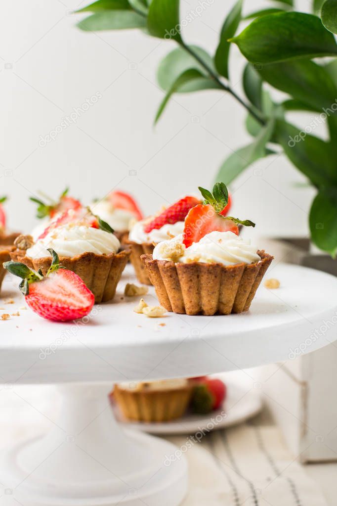 Mini tartlets with cheese cream and fresh strawberries, sprinkled with nuts on a white wooden stand. Food photo