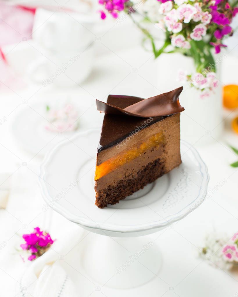 A piece of chocolate mousse cake with apricot on a wooden white background. Fresh apricots and bright flowers