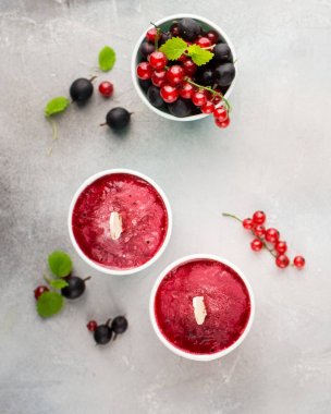 Frozen berry smoothies in bright paper cups on a light gray concrete background with fresh red currant and black gooseberry.place for text. flat lay, top view clipart