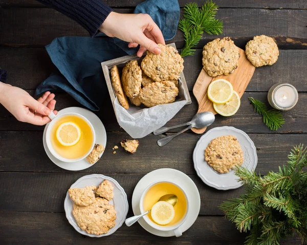 The perfect tasty breakfast. Oatmeal cookies with chocolate in wooden box and green tea with lemon in mugs on a dark background with a Xmas tree. Best useful Cookies. Flat lay, top view