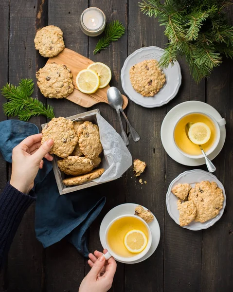 The perfect tasty breakfast. Oatmeal cookies with chocolate in wooden box and green tea with lemon in mugs on a dark background with a Xmas tree. Best useful Cookies. Flat lay, top view