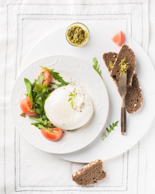 İtalyan peynirli Burrata (mozzarella) taze roka, domates, zeytin yağı, sos pesto ve tahıllı ekmeği beyaz bir tabakta ve ahşap arkaplanda kızartıyorlar.