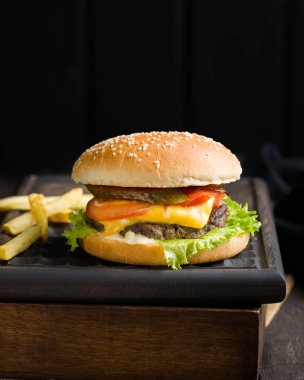 Sığır eti, domates, peynir ve marul, kızarmış patates ve bira ile lezzetli taze lezzetli burger ahşap kesme tahtası koyu arka plan üzerinde servis. Sokak lokanta