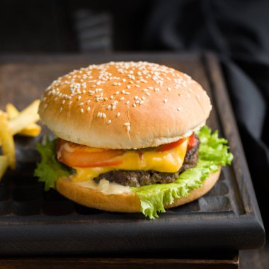 Sığır eti, domates, peynir ve marul, kızarmış patates ve bira ile lezzetli taze lezzetli burger ahşap kesme tahtası koyu arka plan üzerinde servis. Sokak lokanta