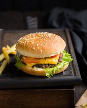 Sığır eti, domates, peynir ve marul, kızarmış patates ve bira ile lezzetli taze lezzetli burger ahşap kesme tahtası koyu arka plan üzerinde servis. Sokak lokanta