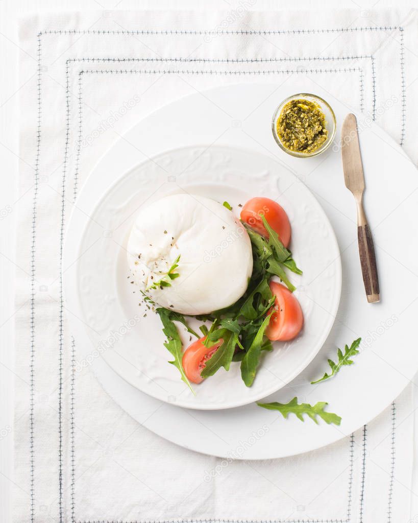 Sliced Italian cheese Burrata (mozzarella) with fresh arugula, tomatoes, olive oil, sauce pesto and whole grain bread toasts on a white plate and a wooden light background.Flat lay, top view