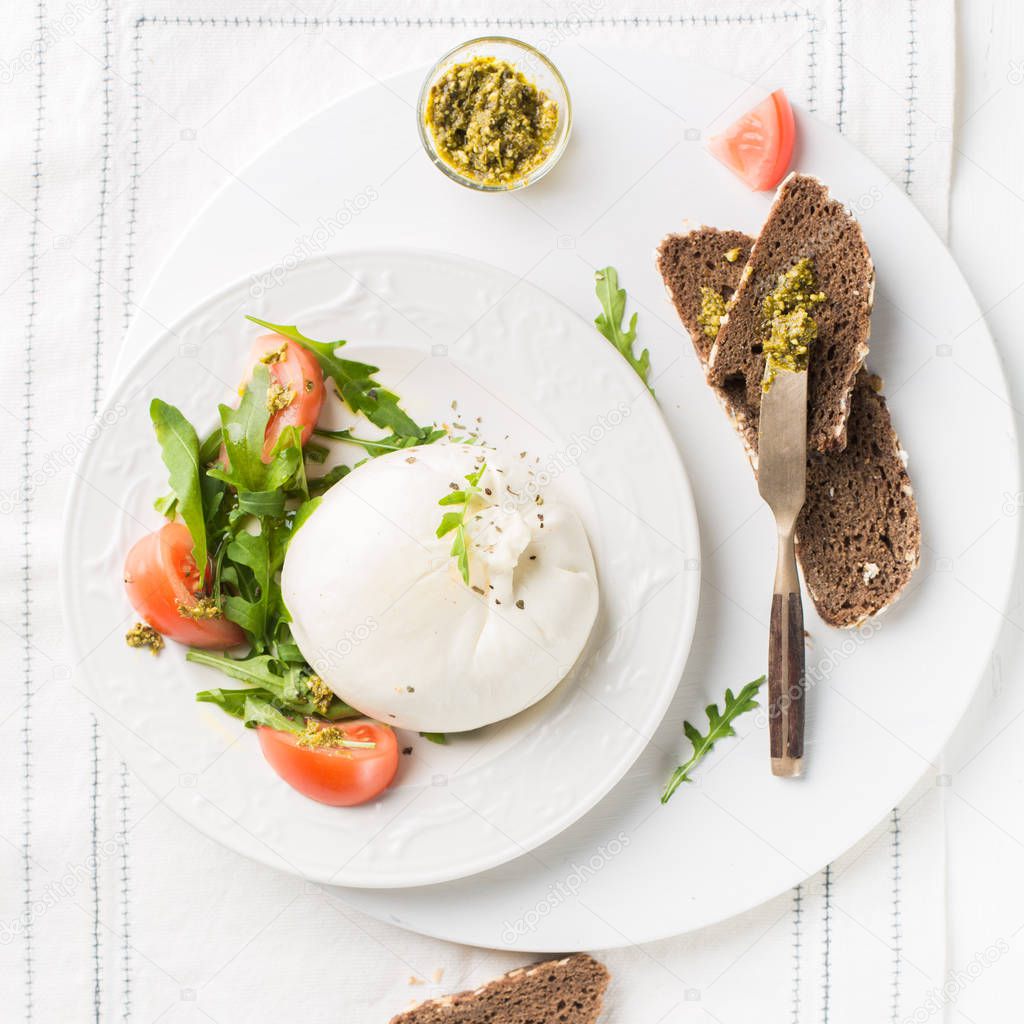 Sliced Italian cheese Burrata (mozzarella) with fresh arugula, tomatoes, olive oil, sauce pesto and whole grain bread toasts on a white plate and a wooden light background.Flat lay, top view