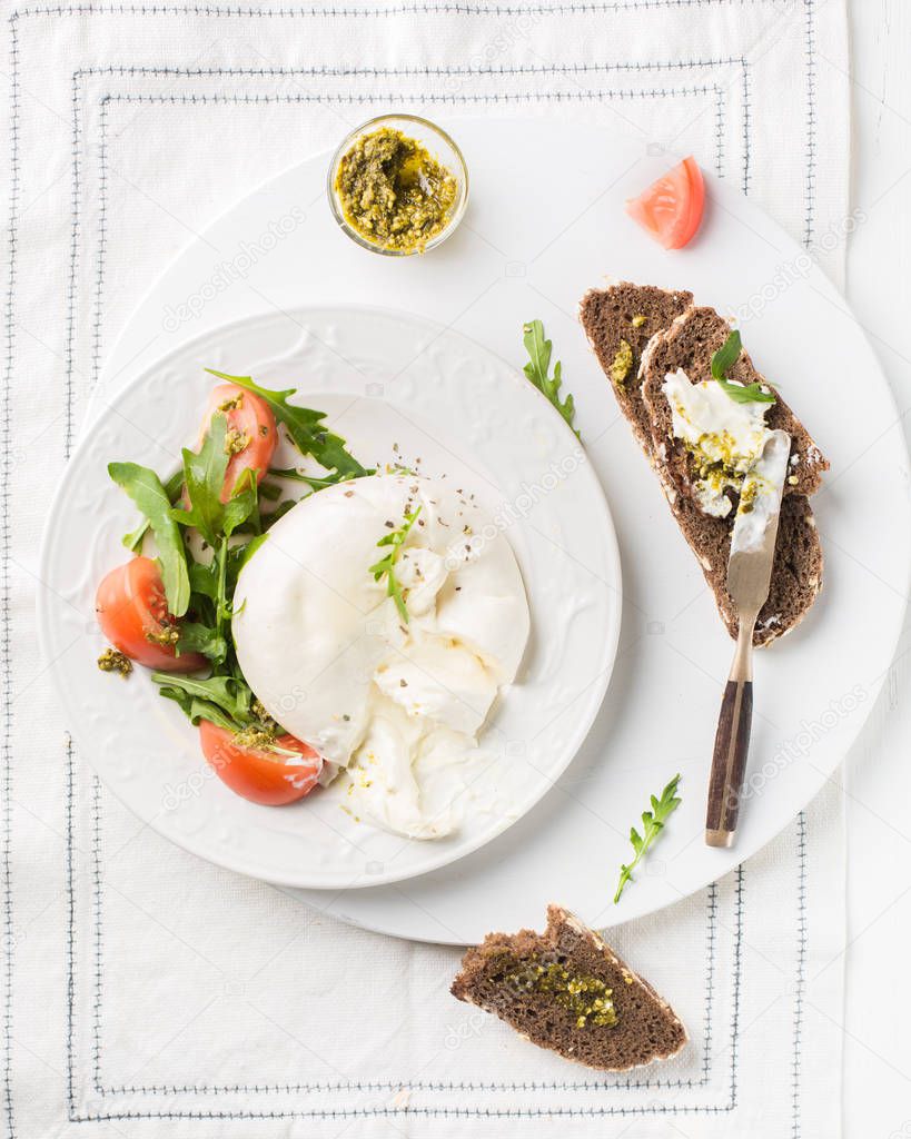 Sliced Italian cheese Burrata (mozzarella) with fresh arugula, tomatoes, olive oil, sauce pesto and whole grain bread toasts on a white plate and a wooden light background.Flat lay, top view