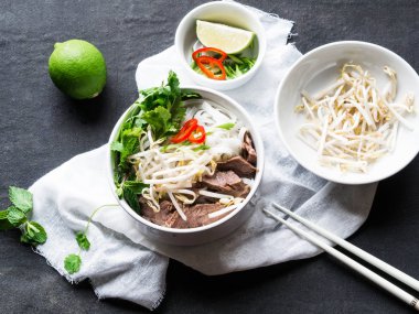 Geleneksel Vietnam çorba pho sığır eti ve pirinç noodle, nane ve Salantro, yeşil soğan, biber, fasulye filizi ve kireç siyah bir zemin üzerine beyaz bir kase. Asya gıda. Üstten Görünüm