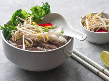 Geleneksel Vietnam çorba-pho sığır eti ve pirinç noodle, nane ve Salantro, yeşil soğan, biber, fasulye filizi ve kireç gri bir arka plan üzerinde beyaz bir kase. Asya gıda