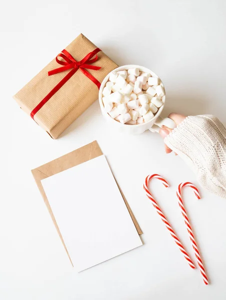 Paper card for letter, envelope, xmas decoration and female hand holds a white mug with hot drink and marshmallows on white background. Top view. Copy space