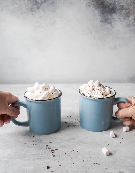 Two blue mugs with hot chocolate, whipped cream, chocolate chips in the hands of uncertain people on gray background