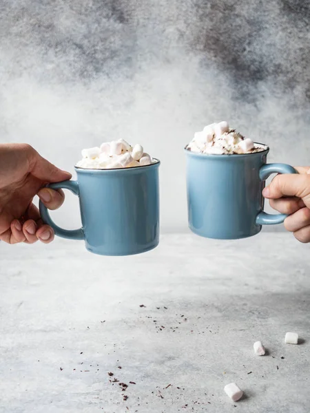 Two blue mugs with hot chocolate, whipped cream, chocolate chips in the hands of uncertain people on a gray background