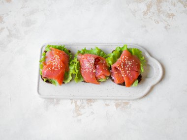 Somon dilimli çavdar ekmeği tostları, gri arka planda beyaz seramik tabakta susam ve marul. Üst görünüm.