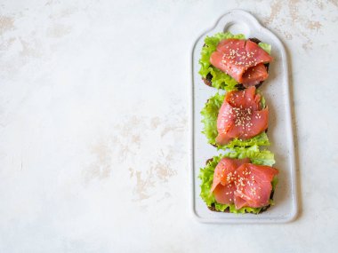Somon dilimli çavdar ekmeği tostları, gri arka planda beyaz seramik tabakta susam ve marul. Üst görünüm.