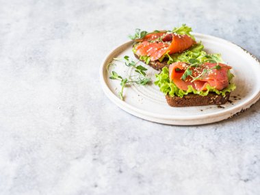 Somon dilimli iki tost, susam, bezelye lahanası ve gri arka planda beyaz seramik tabakta marul. Boşluğu kopyala