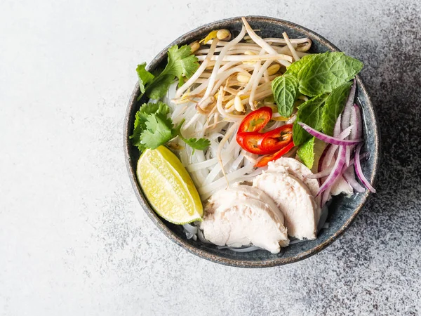 Zuppa tradizionale vietnamita-pho ga in ciotola con pollo e riso tagliatelle, menta e coriandolo, cipolla rossa, peperoncino, germogli di fagioli e lime su fondo grigio. Cibo asiatico . — Foto Stock