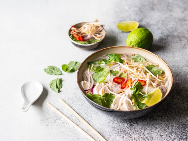 Traditional Vietnamese soup- pho ga in bowl with chicken and rice noodles, mint and cilantro, red onion, chili, bean sprouts and lime on grey background. Asian food. Copy space. — Stock Photo, Image