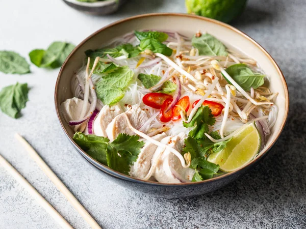 Sopa vietnamita tradicional-pho ga em tigela com macarrão de frango e arroz, hortelã e coentro, cebola vermelha, pimenta, brotos de feijão e limão em fundo cinza. Comida asiática . — Fotografia de Stock