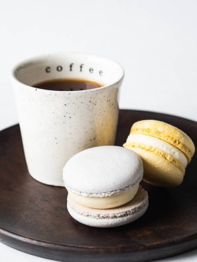 Beyaz seramik fincanlı iki pastel badem kurabiyesi. Ahşap tabakta, beyaz arka planda.