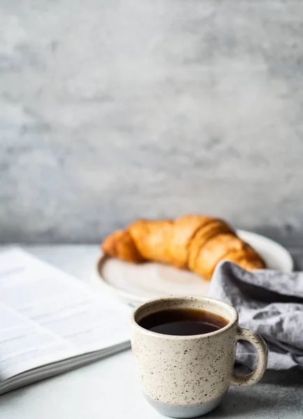 Delicious breakfast - the cup of coffee, croissant and open magazine on the table at the grey background. Copy space.