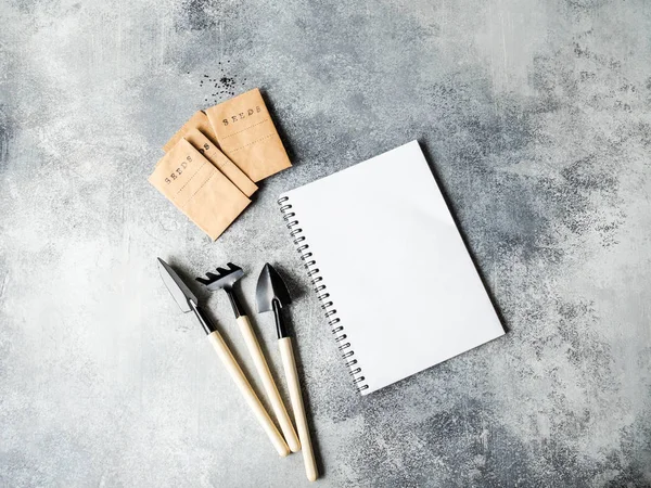 Open white notebook for text, garden tools and seeds in paper bags on a gray background. Top view. Garden concept