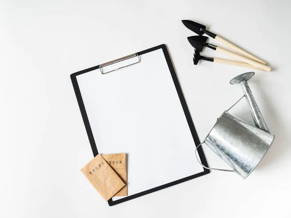 Clipboard and paper for text, garden tools and seeds in paper bags on white background. Garden concept. Top view.