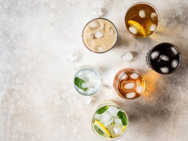 Buzlu bardaklarda çeşitli ferahlatıcı içeceklerin olduğu düz bir yer. Elma suyu, kola, ev yapımı limonata, buzlu kahve, buzlu çay ve bej arka planda köpüklü su. Üst görünüm