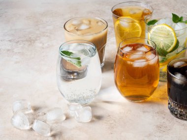 Buzlu bardaklarda çeşitli ferahlatıcı içecekler. Elma suyu, kola, ev yapımı limonata, buzlu kahve, buzlu meyve çayı ve bej arka planda köpüklü su. Boşluğu kopyala